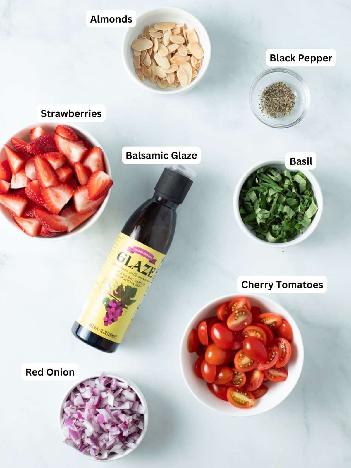 Strawberry tomato salad ingredients in small bowls: almonds, strawberries,  black pepper, basil, balsamic glaze, cherry tomatoes, red onion.