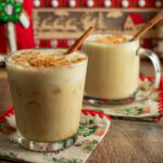 Glass filled with vegan eggnog, and topped with cinnamon and a cinnamon stick, with another mug behind it filled with vegan eggnog.