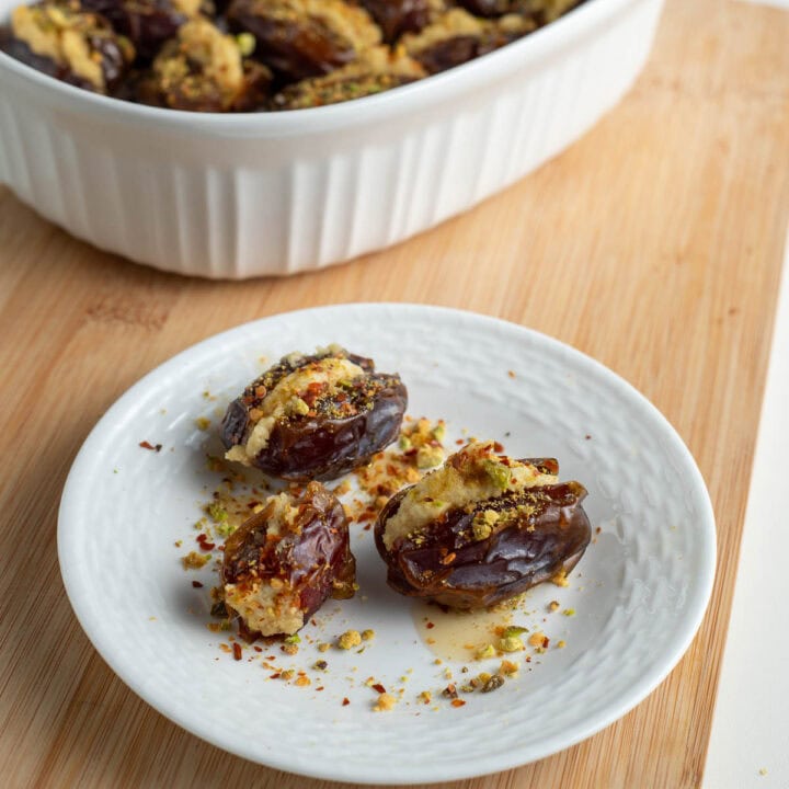 Vegan cashew stuffed dates on a small white plate topped with pistachios and red pepper flakes.